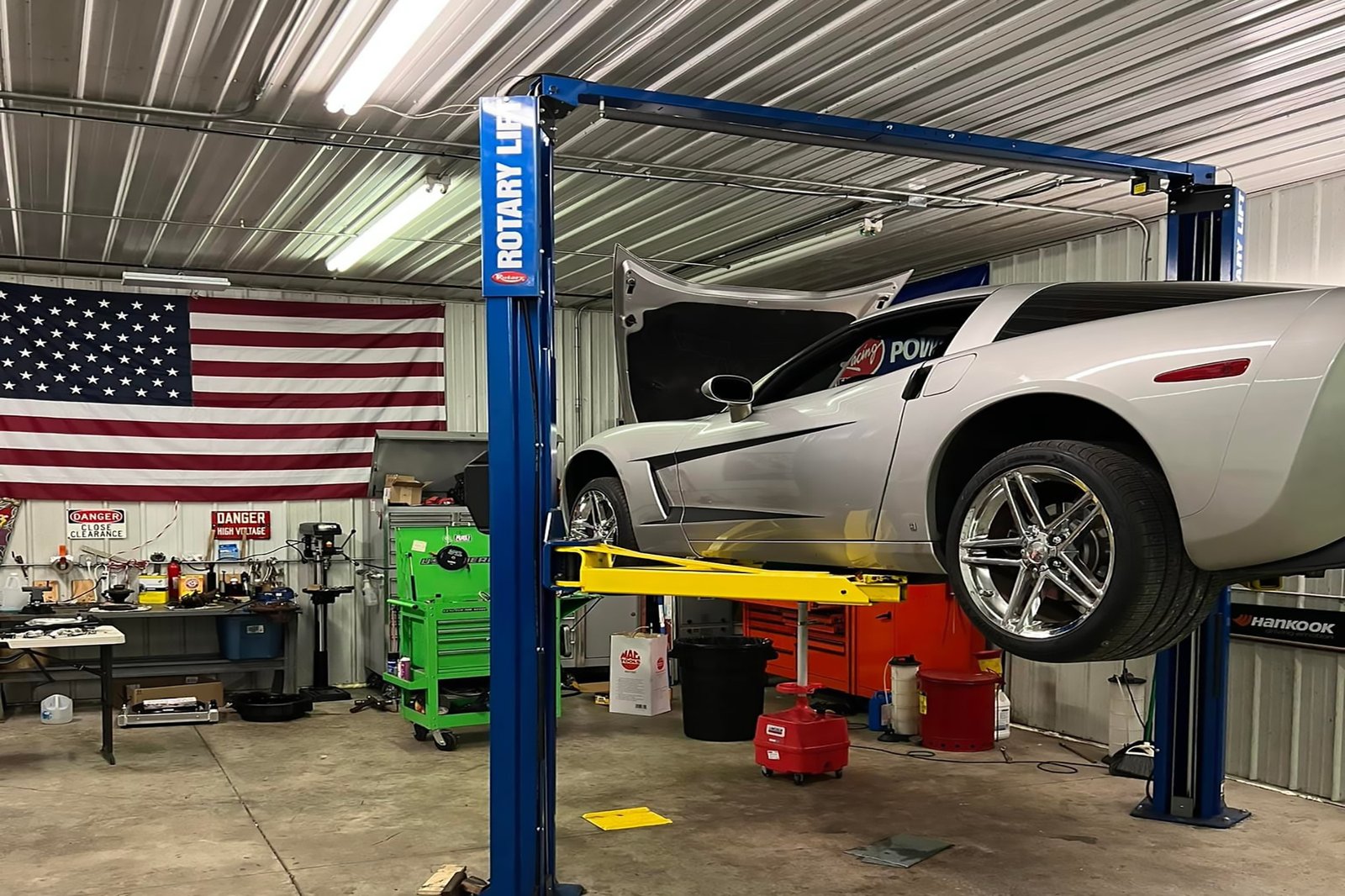A silver Chevrolet Corvette raised on a professional lift for performance tuning at Grayless Automotive in Clinton County, featuring a clean workshop with a prominent American flag.