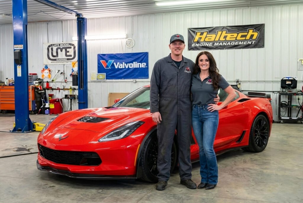Owners Braden and Tabby Grayless standing in the Grayless Automotive workshop in front of a silver Chevrolet Corvette. Braden is wearing mechanic coveralls and Tabby is in a branded company polo.